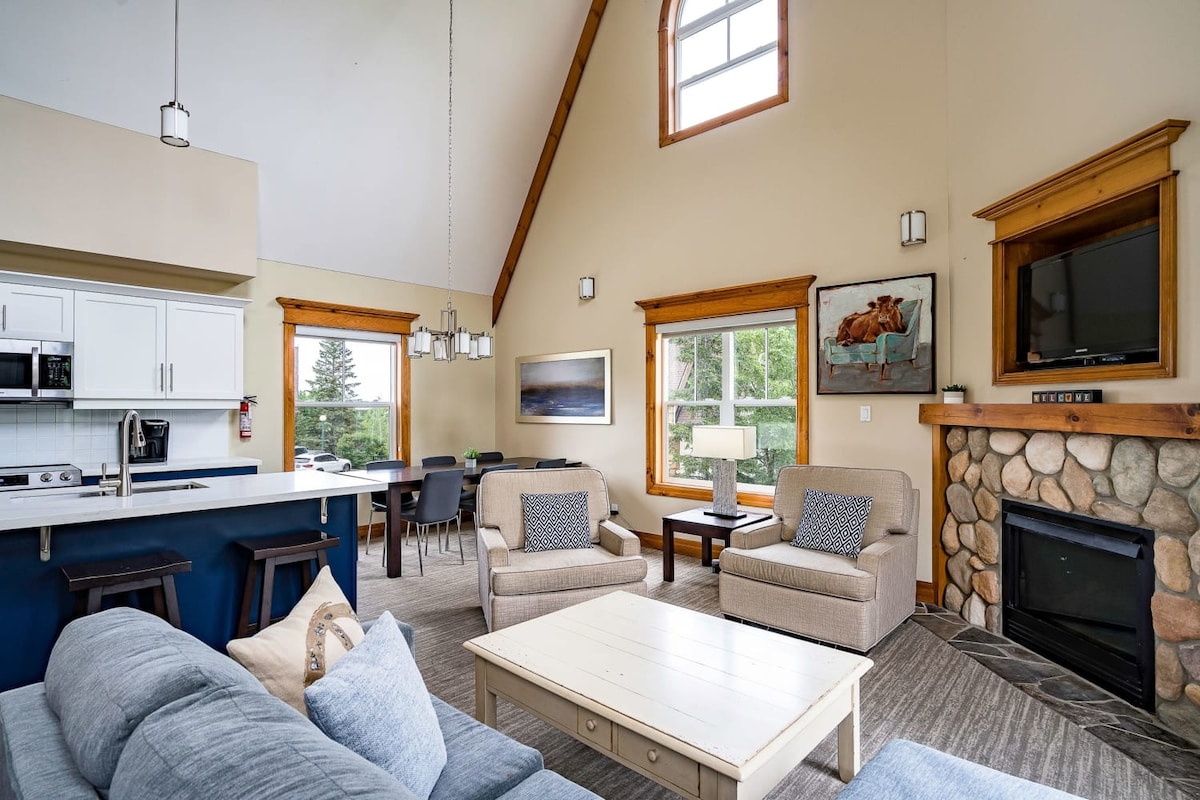 The open-concept living area features cathedral ceilings and large windows that provide natural light. A cozy fireplace is surrounded by comfortable seating, including two armchairs and a spacious sofa, with a coffee table in the center. The kitchen area, equipped with modern appliances, is visible in the background.