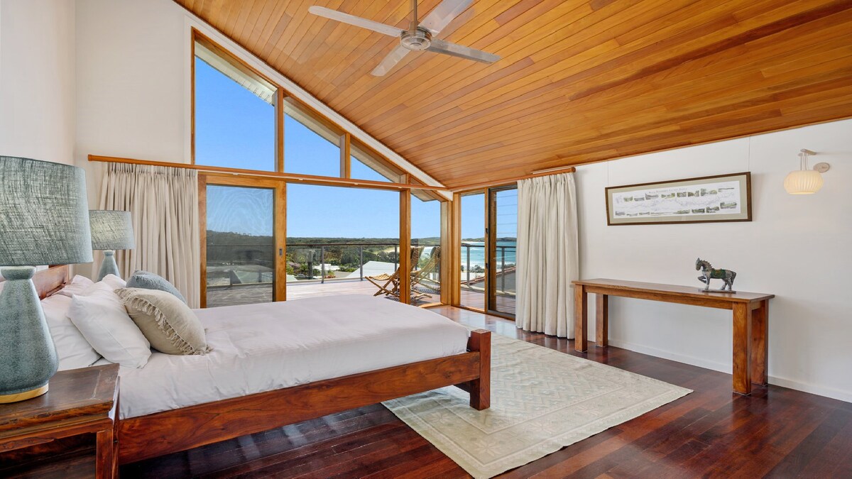 A spacious bedroom features a wooden bed frame with soft linens and decorative pillows. Natural light floods the room through large windows with stunning views. A wooden table adds a touch of elegance, while wooden ceiling beams enhance the airy feel.