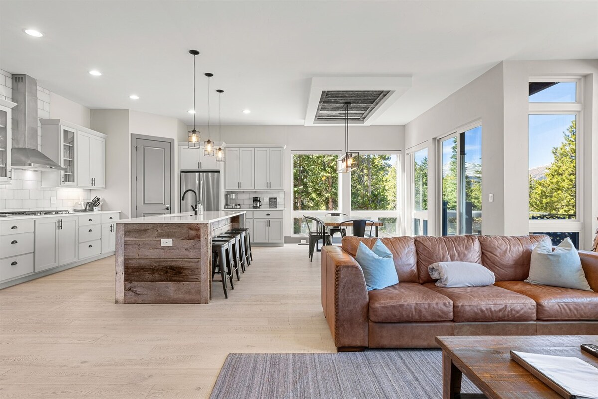 An open living space is showcased, featuring a contemporary kitchen with white cabinetry and a large island. A cozy leather sofa is arranged in the seating area, while large windows frame scenic views of the surroundings, allowing natural light to brighten the room.