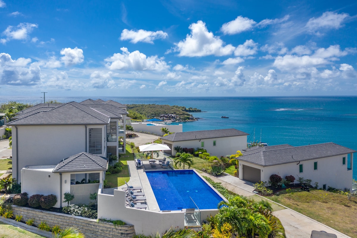 An aerial view captures the luxury residences along the southern coast of Grenada, showcasing a spacious community pool surrounded by lounge chairs. Lush greenery complements the landscape, and the sparkling ocean can be seen in the background under a bright blue sky adorned with fluffy clouds.