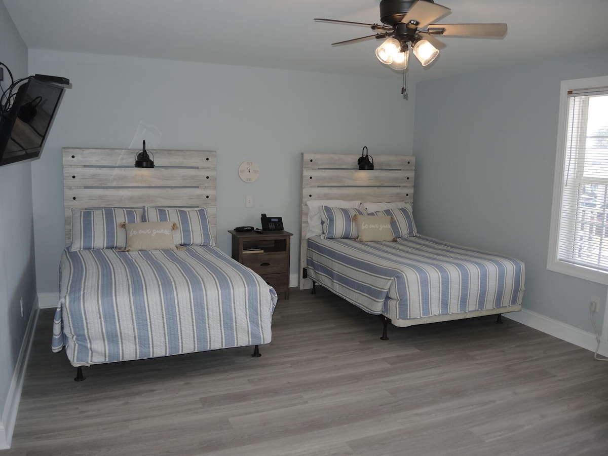 A welcoming studio room features two spacious beds with striped blue and white bedding. A wooden accent wall complements the neutral tones, while a ceiling fan provides airflow. A small nightstand and a wall-mounted television are also visible.