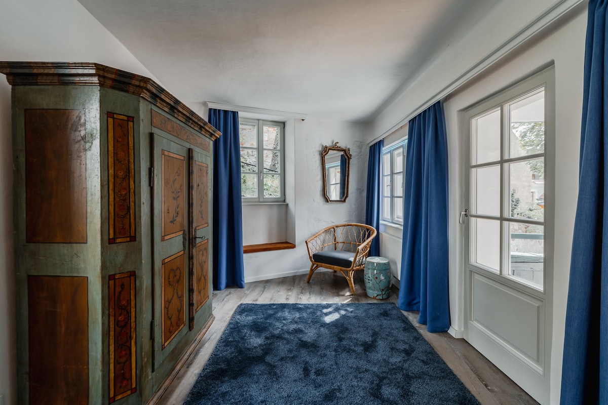 A thoughtfully arranged entryway features a vintage cabinet with intricate artwork, complemented by a rattan chair. Natural light filters through two windows, accented by deep blue curtains. A round mirror adds depth to the space, while a small table near the door offers functionality.