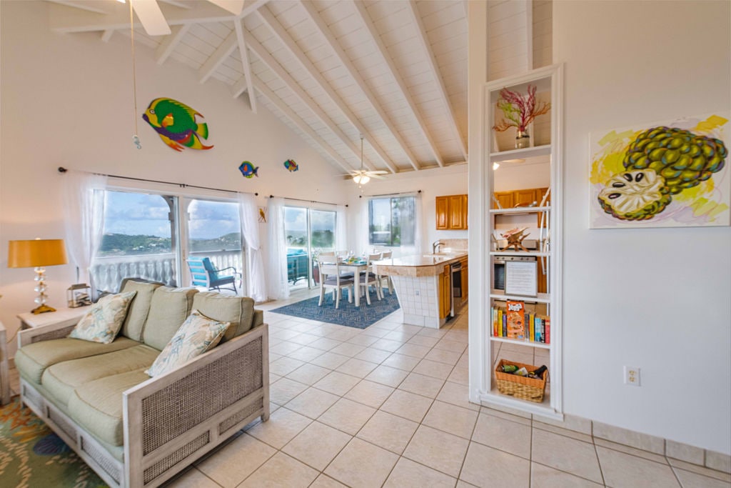 An open living space features a light-filled area with vaulted ceilings. A comfortable sofa is positioned near expansive windows overlooking scenic views. A dining area and well-equipped kitchen are visible, with tasteful decor on light walls and colorful accents adding vibrancy.