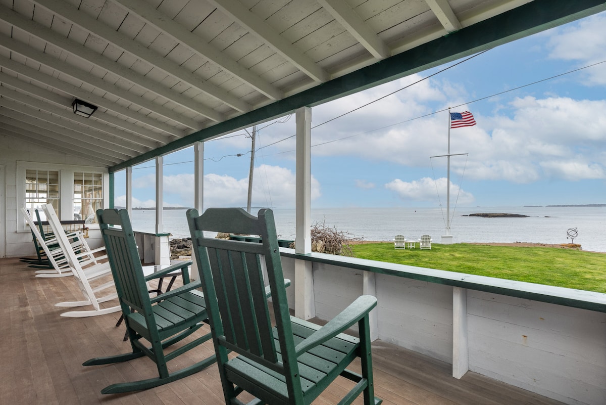 A covered porch features several rocking chairs, providing a comfortable space to enjoy views of the water and distant islands. The scene is complemented by an American flag gently waving in the breeze and a well-maintained lawn in the foreground.