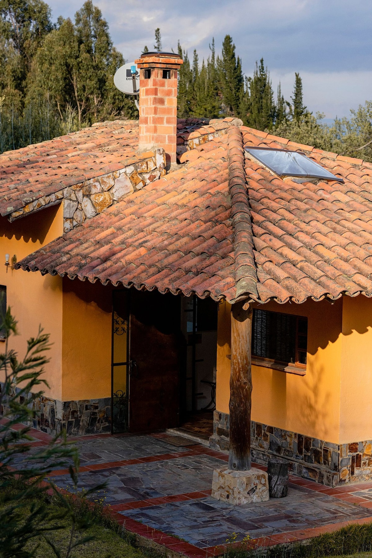 A charming cottage exterior features a terracotta roof with a skylight, complemented by a stone chimney. The warm orange walls and stone accents provide a welcoming feel, while the natural surroundings of trees are visible in the background.