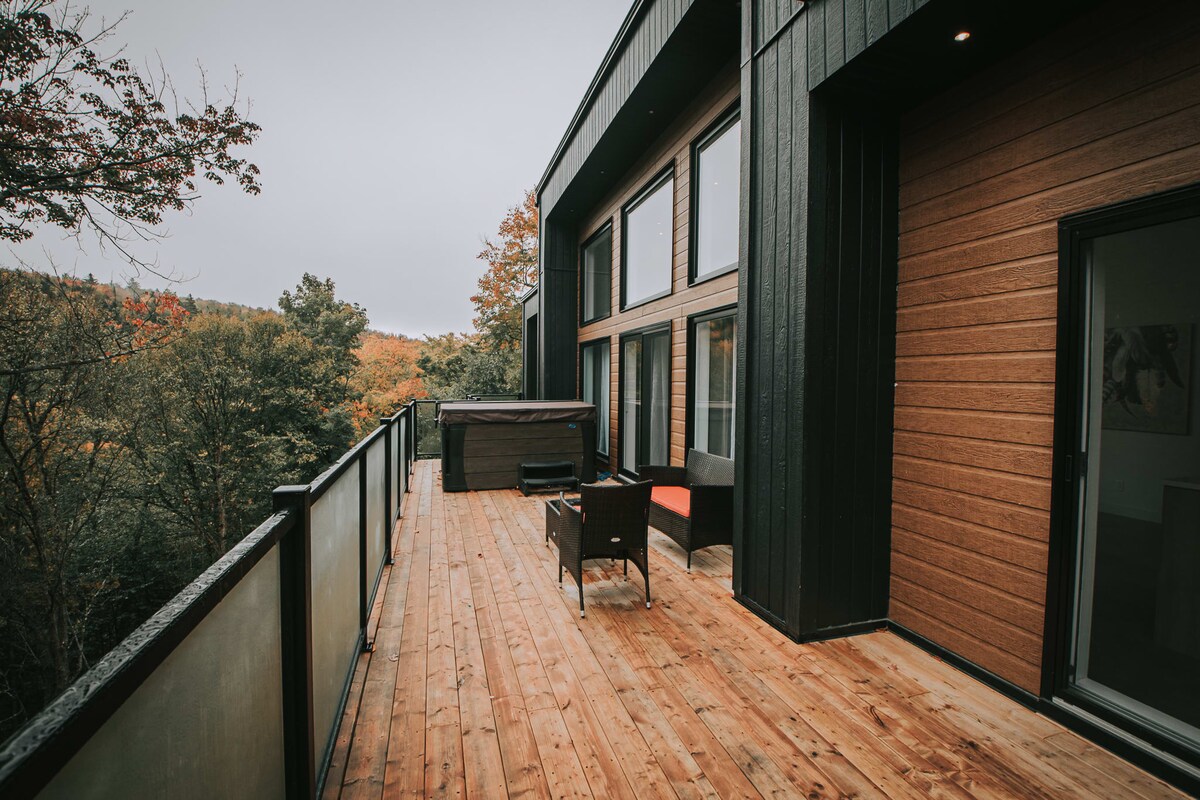 A spacious wooden deck features a hot tub and comfortable seating, with views of surrounding trees adorned in autumn colors. Large windows of the chalet reflect the natural landscape, offering a seamless connection to the outdoors.