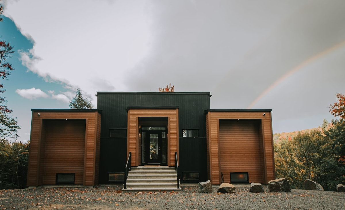 The modern chalet features a striking façade with a combination of dark and light wood tones. Two distinct sections flank a central entrance, leading to a set of steps that invite guests to enter. Natural surroundings enhance the rural charm.