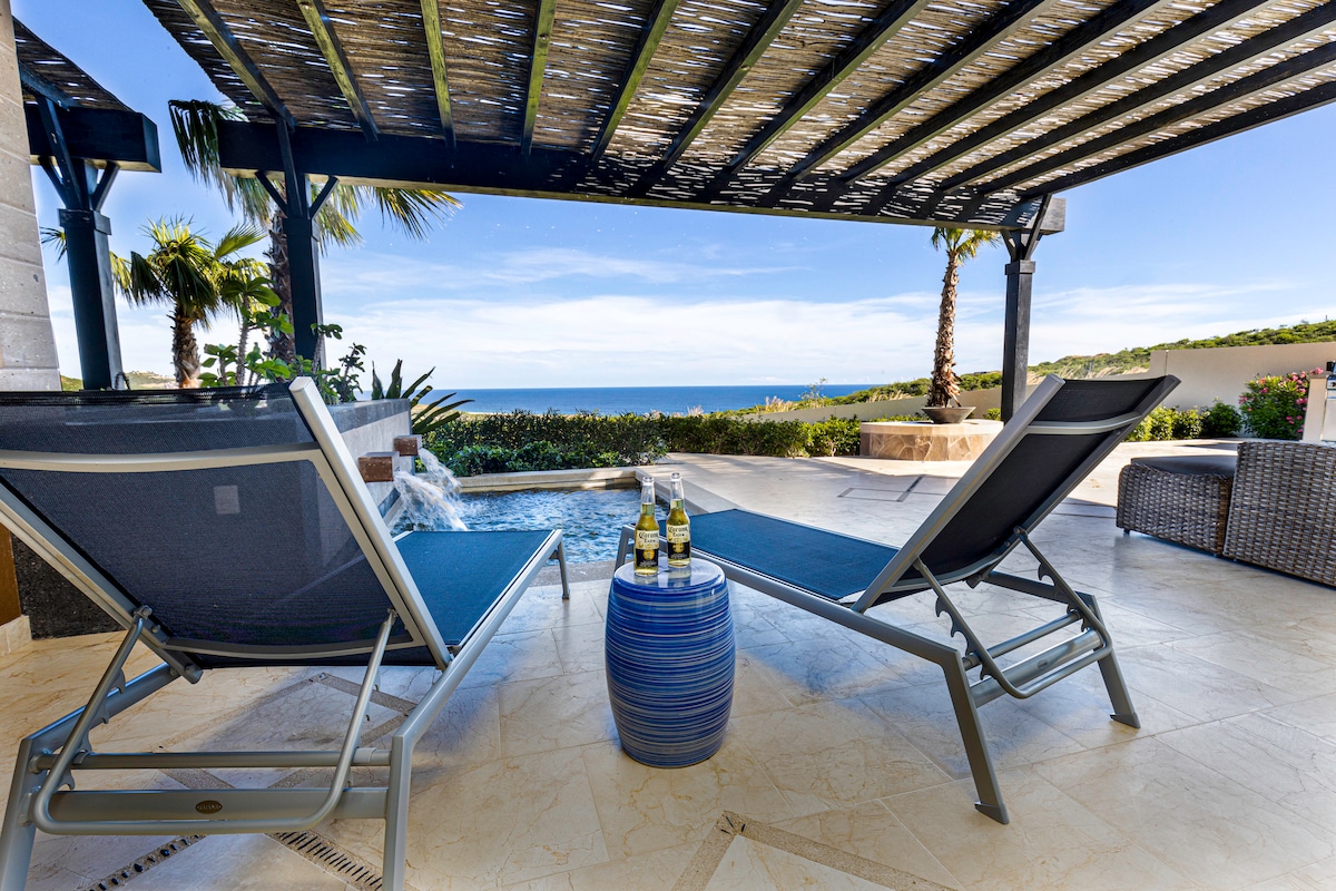 A private terrace features lounge chairs positioned beside a small dipping pool, with ocean views visible in the background. A small table holds two bottles, adding a relaxed touch to the outdoor space shaded by a pergola.