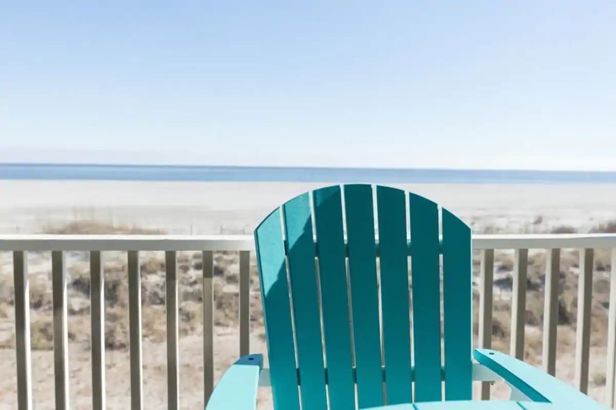 A bright blue Adirondack chair is positioned on a balcony, overlooking a sandy beach and tranquil ocean waters. The wide expanse of the beach stretches into the distance, with soft, natural hues blending under a clear sky.