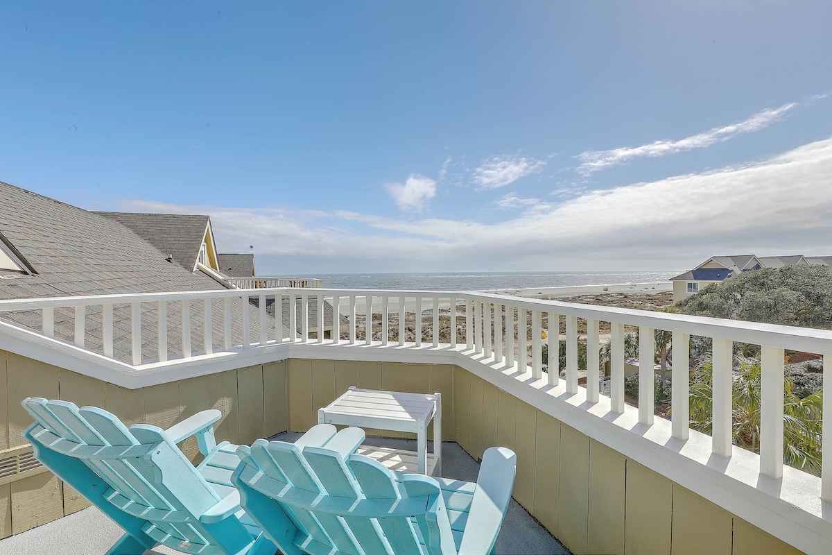 A rooftop area displays two light blue adirondack chairs positioned around a small white table, offering an unobstructed view of the ocean. The scene is complemented by a clear blue sky and soft clouds, highlighting the serene coastal atmosphere.