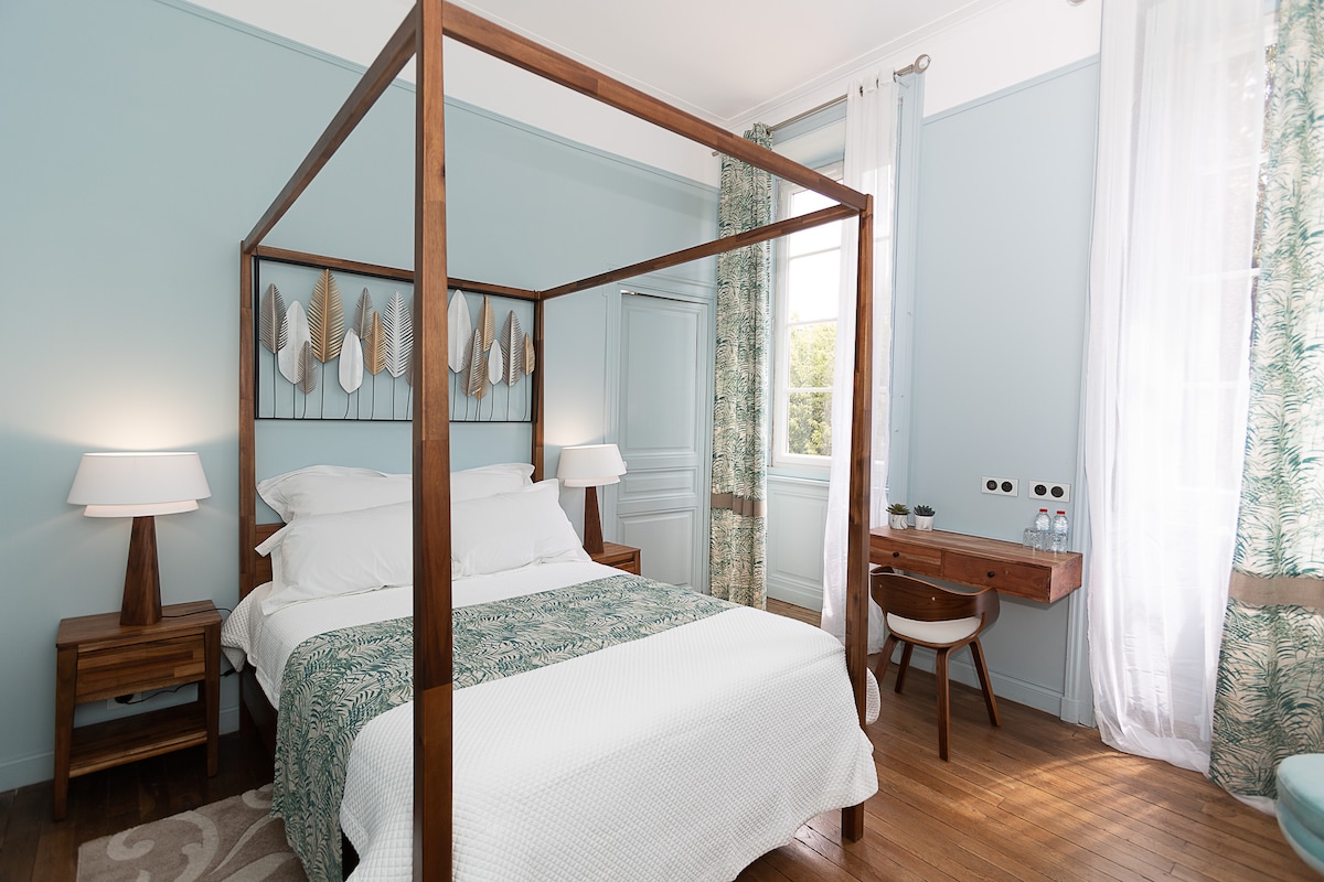 A spacious bedroom features a four-poster bed adorned with a white quilt. Light green walls complement the natural decor above the headboard. Bedside tables with lamps provide soft illumination, while a wooden desk sits by a window dressed in sheer curtains, enhancing the airy feel of the room.