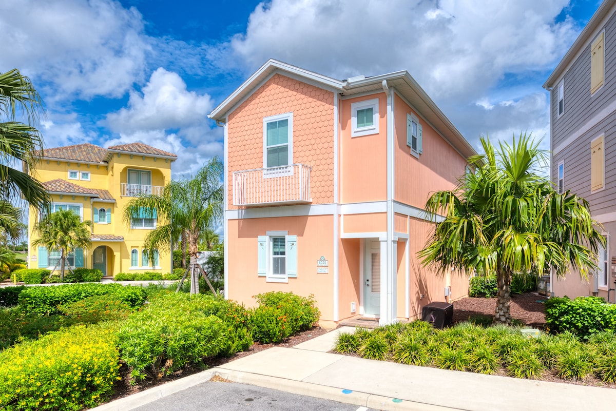 A charming cottage in soft peach color is surrounded by lush greenery and vibrant landscaping. The exterior features a small balcony on the upper level, while palm trees add to the tropical atmosphere. The pathway leads to a well-maintained entrance, showcasing the inviting nature of the property.