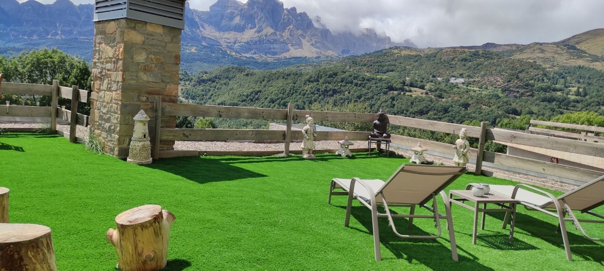 A spacious terrace is presented, featuring comfortable lounge chairs placed on a vibrant green lawn. In the background, stunning mountain views are visible, complemented by a wooden fence. A stone feature and a barbecue setup are also included, creating a welcoming outdoor space.