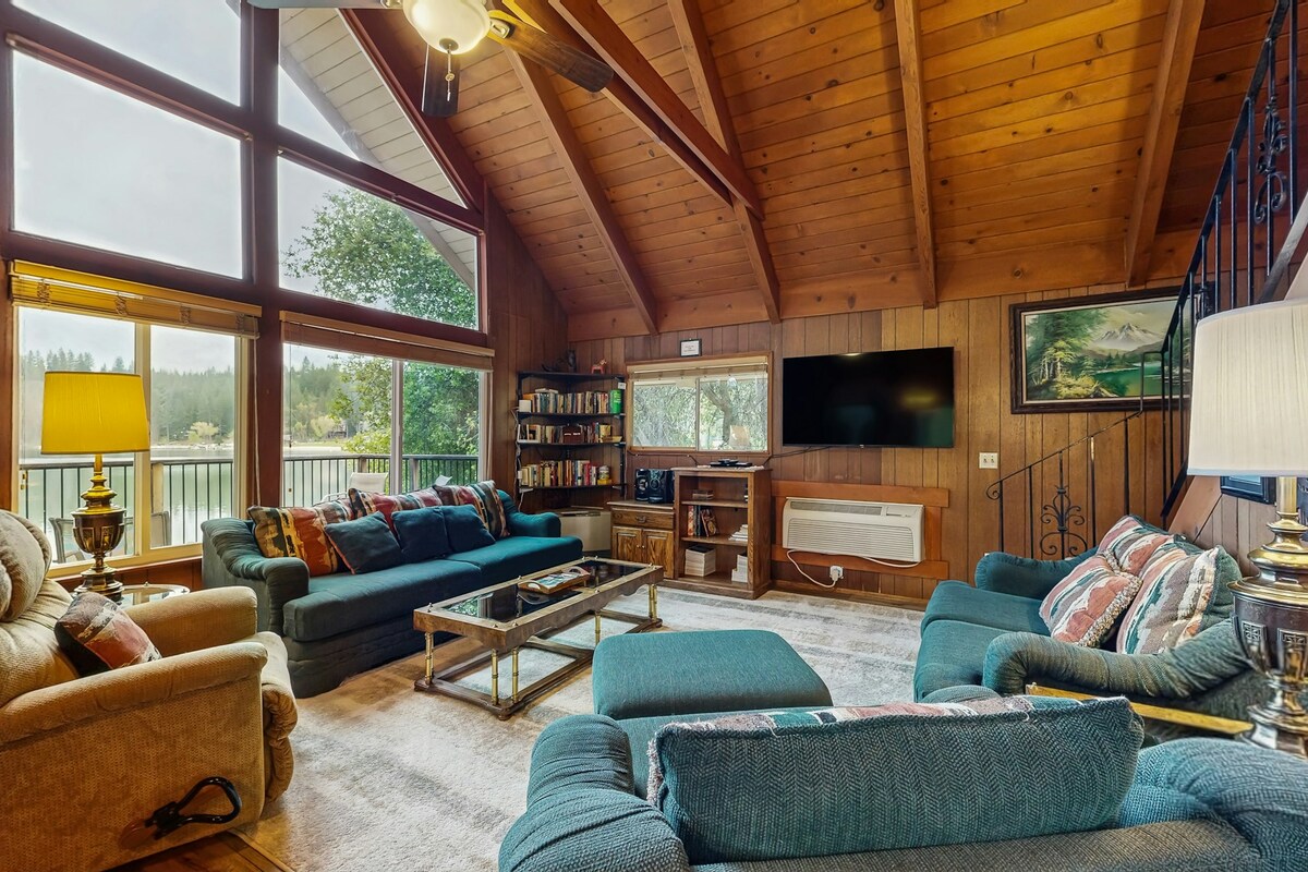 A spacious living room is featured with high wooden ceilings and large windows that provide ample natural light. Comfortable seating includes a teal sofa and an armchair. A coffee table occupies the center, while a bookshelf and media console are positioned against the walls.
