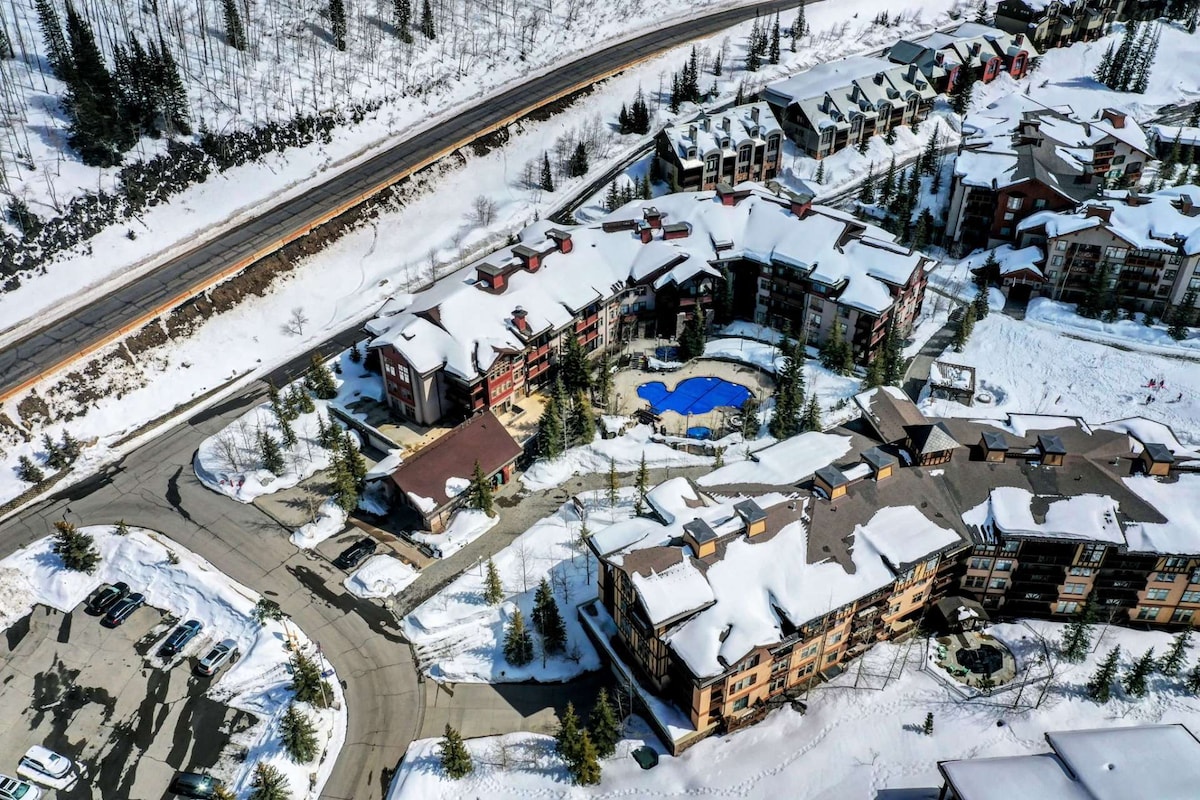 An aerial view captures the snow-covered Solitude Mountain Resort, showcasing the layout of the buildings and the outdoor community pool. The surrounding trees and clear roads highlight the winter landscape, with parked vehicles lining the designated parking areas.