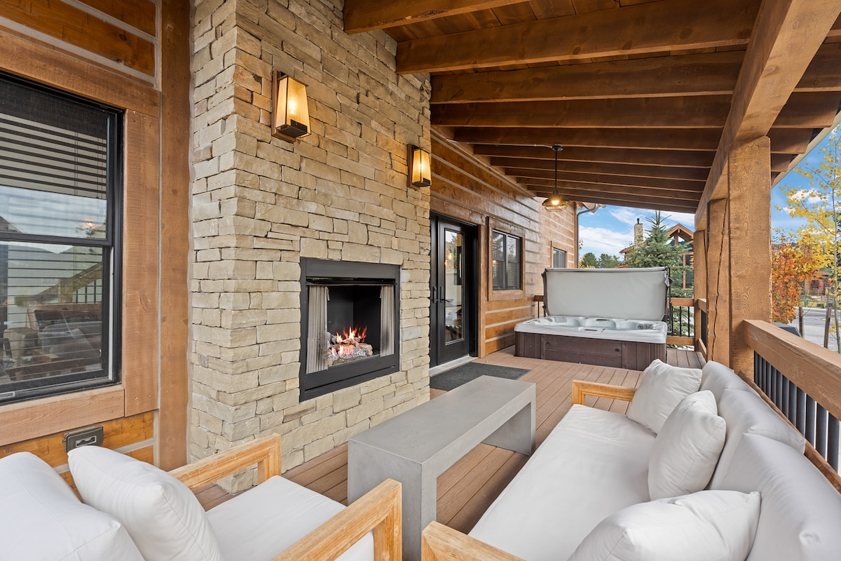 A welcoming covered patio features comfortable seating with plush white cushions arranged around a stone coffee table. A stone fireplace provides warmth, while a hot tub is situated nearby. The area is framed by wooden beams and offers views of the outdoors.