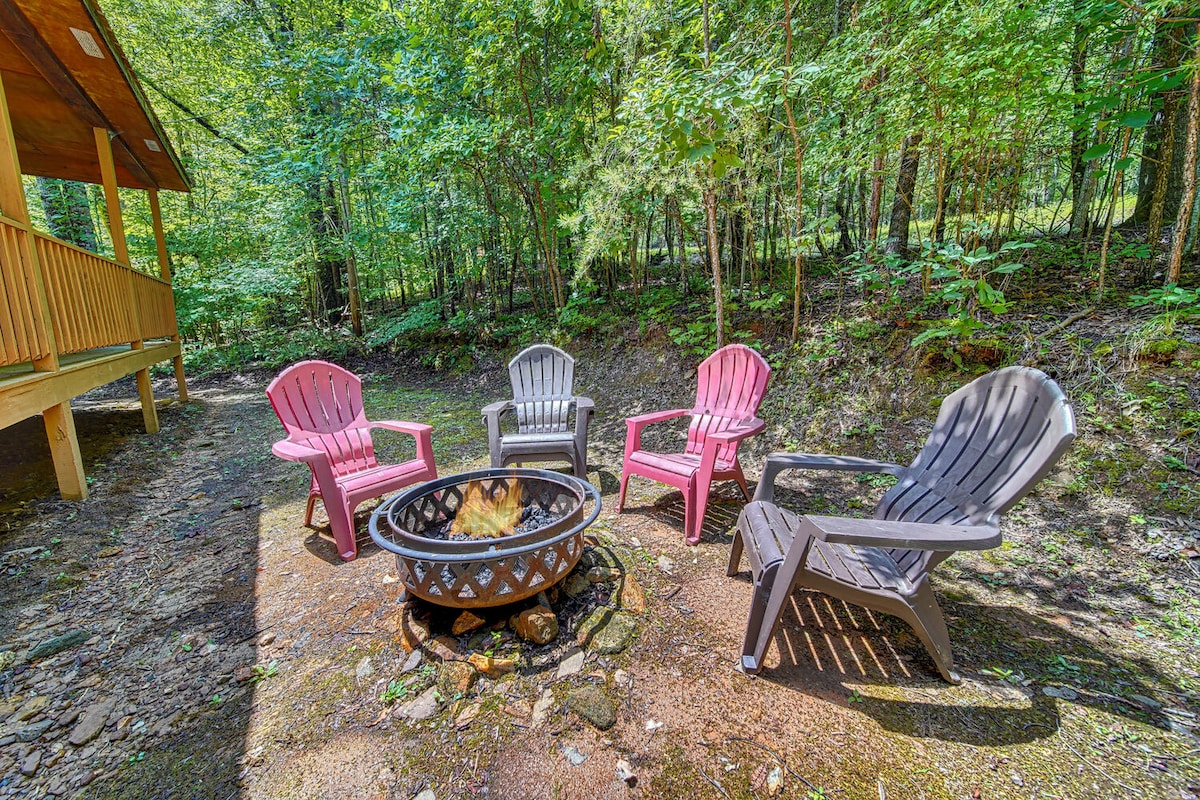 A firepit is surrounded by four colorful chairs—two in pink and two in gray—set in a natural clearing. The scene is framed by lush greenery, creating a peaceful outdoor space for gatherings and relaxation.