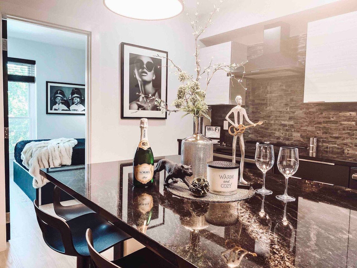 A modern kitchen island, featuring a dark granite countertop, is adorned with a champagne bottle and two glasses. Black cabinetry and a stone backsplash provide a sleek backdrop. Framed art on the walls adds a stylish touch, while a cozy bedroom is visible in the background.