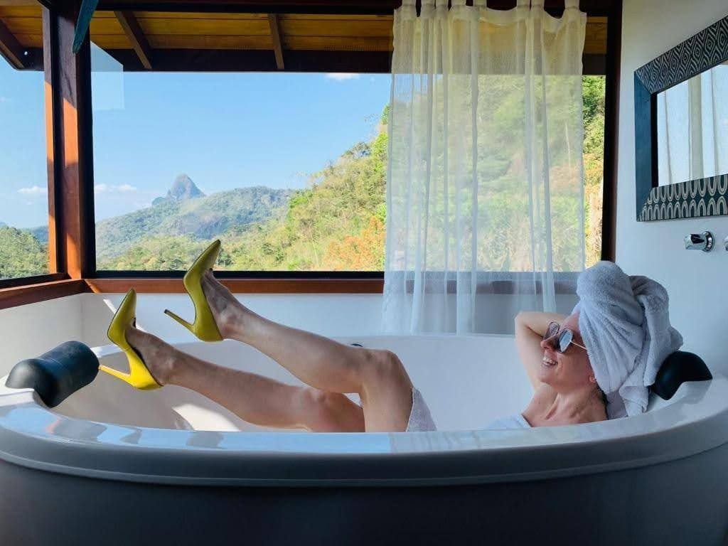 A sunlit bathroom features a spacious bathtub positioned beside large windows, offering an expansive view of the surrounding mountains. Vibrant yellow high heels are visible on the edge of the tub, while soft, sheer curtains gently diffuse natural light into the space.