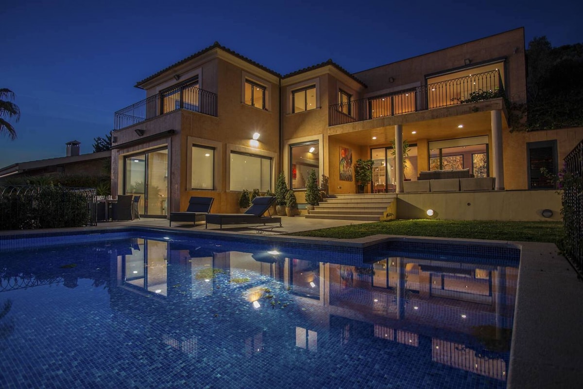 The villa is illuminated at night, with a private pool reflecting the building's lights. Spacious outdoor lounging areas are visible, along with large glass doors that provide a view of the interior. Lush greenery surrounds the property, enhancing the tranquil setting.