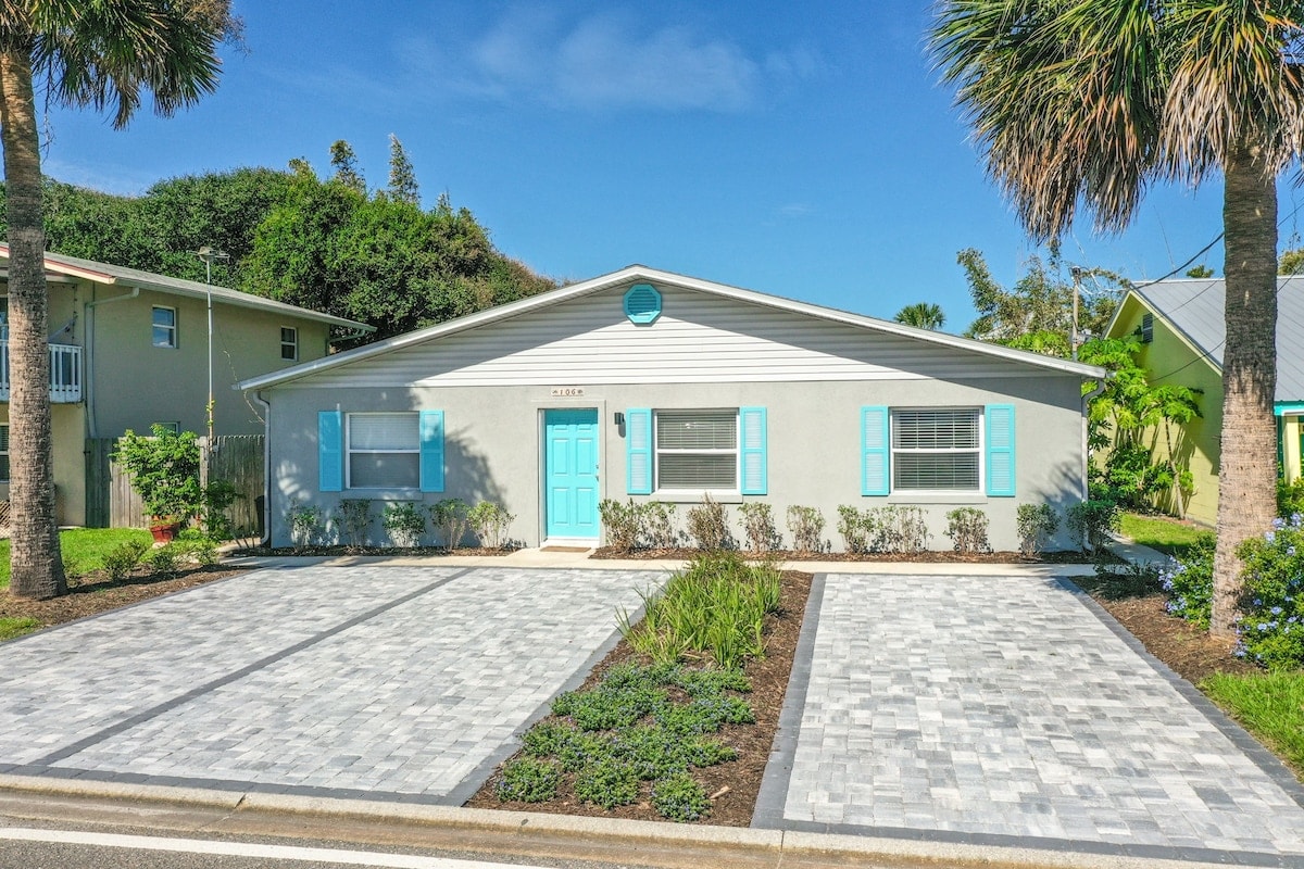 A charming single-story home showcases a light exterior complemented by blue accents. The manicured landscape features shrubs along the front, and a large driveway made of interlocking pavers leads to the welcoming entrance.