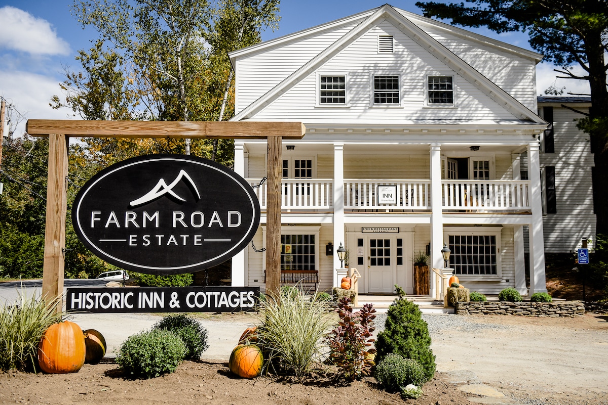 The entrance of Farm Road Estate features a large wooden sign displaying 'Historic Inn & Cottages.' The building's exterior displays classic architectural elements with white siding and multiple balconies. Seasonal pumpkins and plants are positioned in the foreground, enhancing the inviting atmosphere.