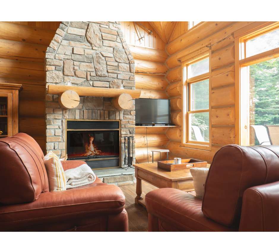 A cozy living room features a large stone fireplace with a wood mantel as its focal point. Comfortable leather sofas are positioned around a wooden coffee table. Natural light enters through large windows, highlighting the warm wooden log walls and inviting atmosphere.