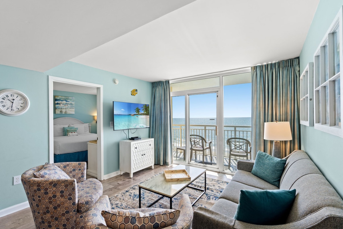 A coastal-themed living area features a comfortable grey sofa and an accent chair with a unique pattern. A glass coffee table sits in the center, and a flat-screen TV is mounted on the wall. Large sliding glass doors lead to a balcony showcasing ocean views.