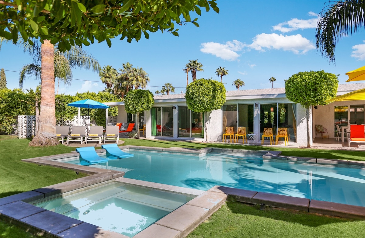 A resort-style backyard features a private pool with a connected hot tub, surrounded by manicured grass. Colorful lounge chairs and shaded seating areas are placed around the pool. Tall hedges and palm trees provide privacy, while bright blue skies and scattered clouds create a vibrant setting.