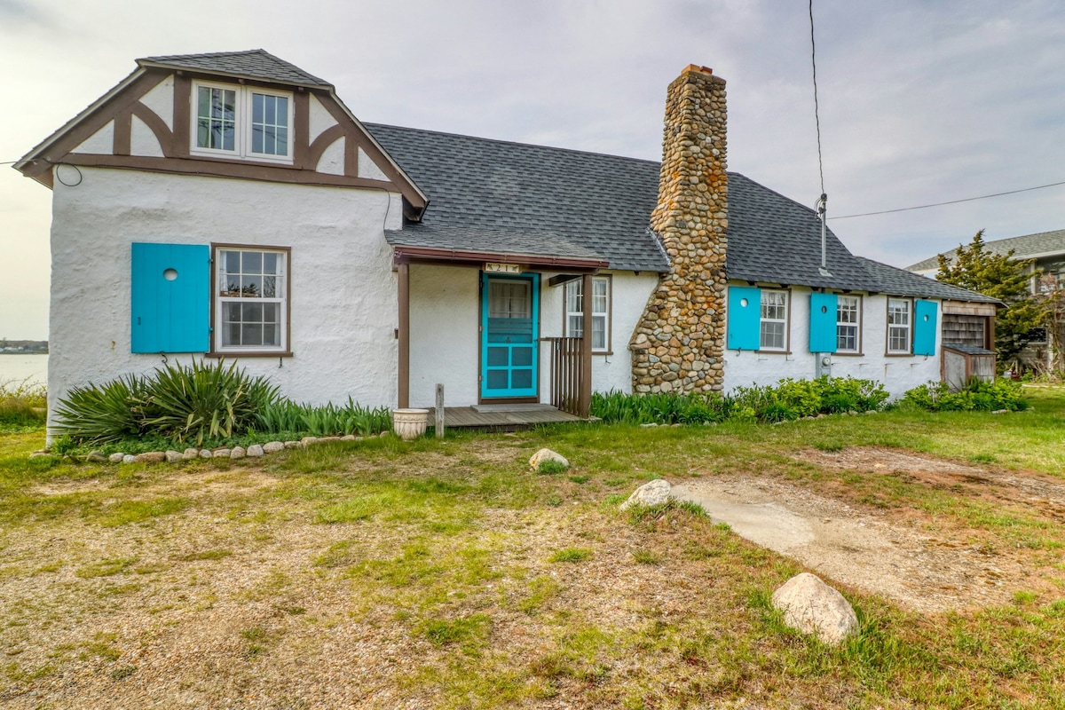 A charming beach house features a stone chimney and bright blue window shutters. The exterior is painted in a light hue, complemented by lush greenery in the front yard. A gravel path leads to the entrance, showcasing the inviting coastal design.