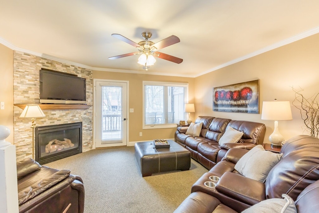 The living room features comfortable leather seating arranged around a coffee table. A wall-mounted television is positioned above a gas fireplace, with large windows allowing natural light. Soft lighting from table lamps creates a cozy ambiance.