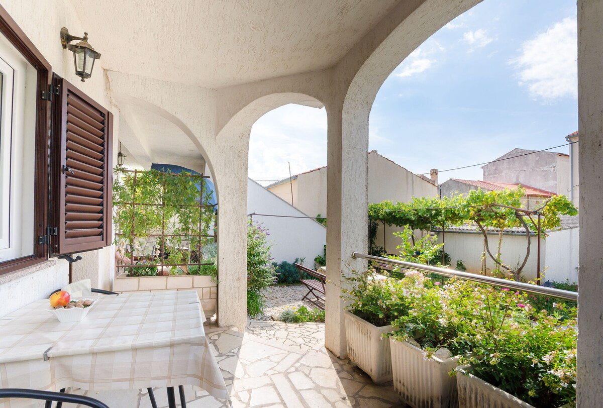 A spacious terrace is framed by arches, featuring a table set with a bowl of fruit. Lush greenery and flowering plants create a natural barrier, while a stone pathway leads to an inviting outdoor area. Sunlight filters through, enhancing the tranquil atmosphere.