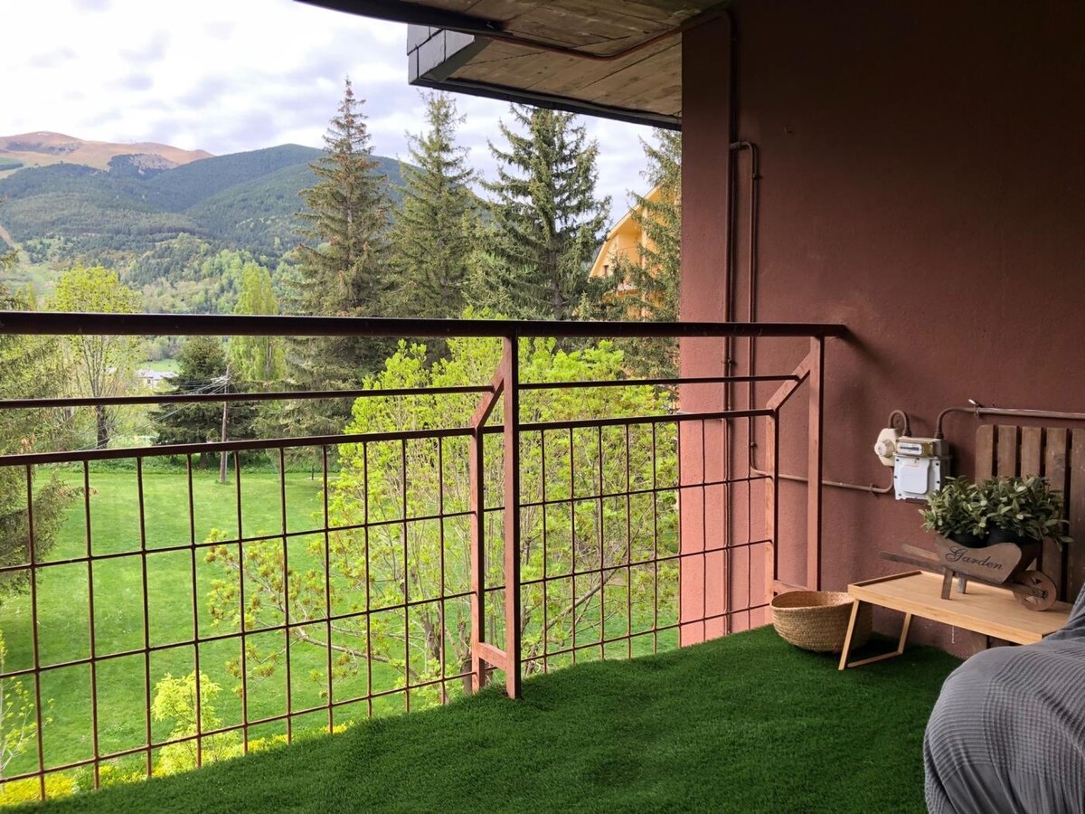A private terrace features a low railing overlooking verdant green fields, framed by trees. The view includes distant mountains, visible under a partly cloudy sky. A small wooden table is accompanied by a woven basket, enhancing the serene outdoor space.