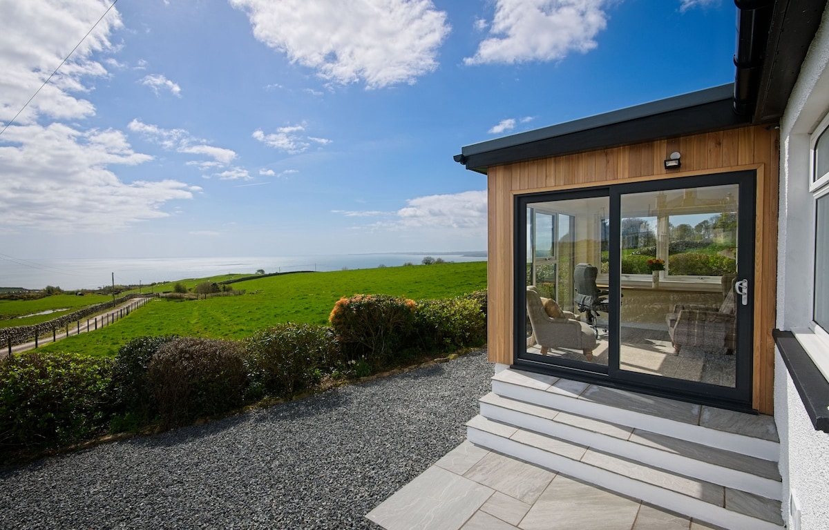 A view of the cottage's exterior features a large glass door leading into a sunlit room. Surrounding greenery and a gravel path provide a pleasant contrast to the modern design. The coastline can be seen in the distance, framed by a clear blue sky with scattered clouds.