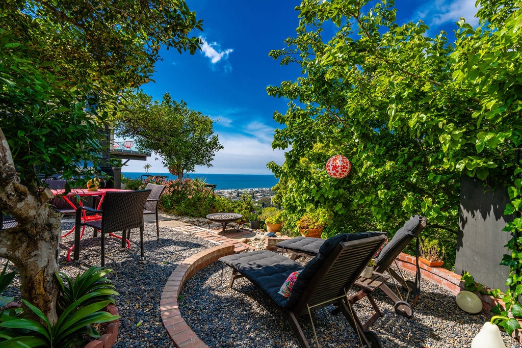 A serene outdoor space features lounge chairs positioned around a circular area, surrounded by lush greenery. A small dining table with chairs is visible amidst the vibrant landscape, offering views of the ocean in the distance under a clear blue sky.