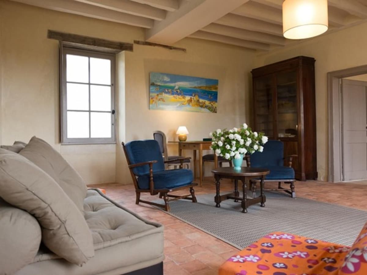 A spacious living area features a comfortable sofa and two blue armchairs arranged around a round coffee table. A vase of flowers sits at the center, while a large window allows daylight to illuminate the room. A painting depicting a serene landscape adorns the wall.