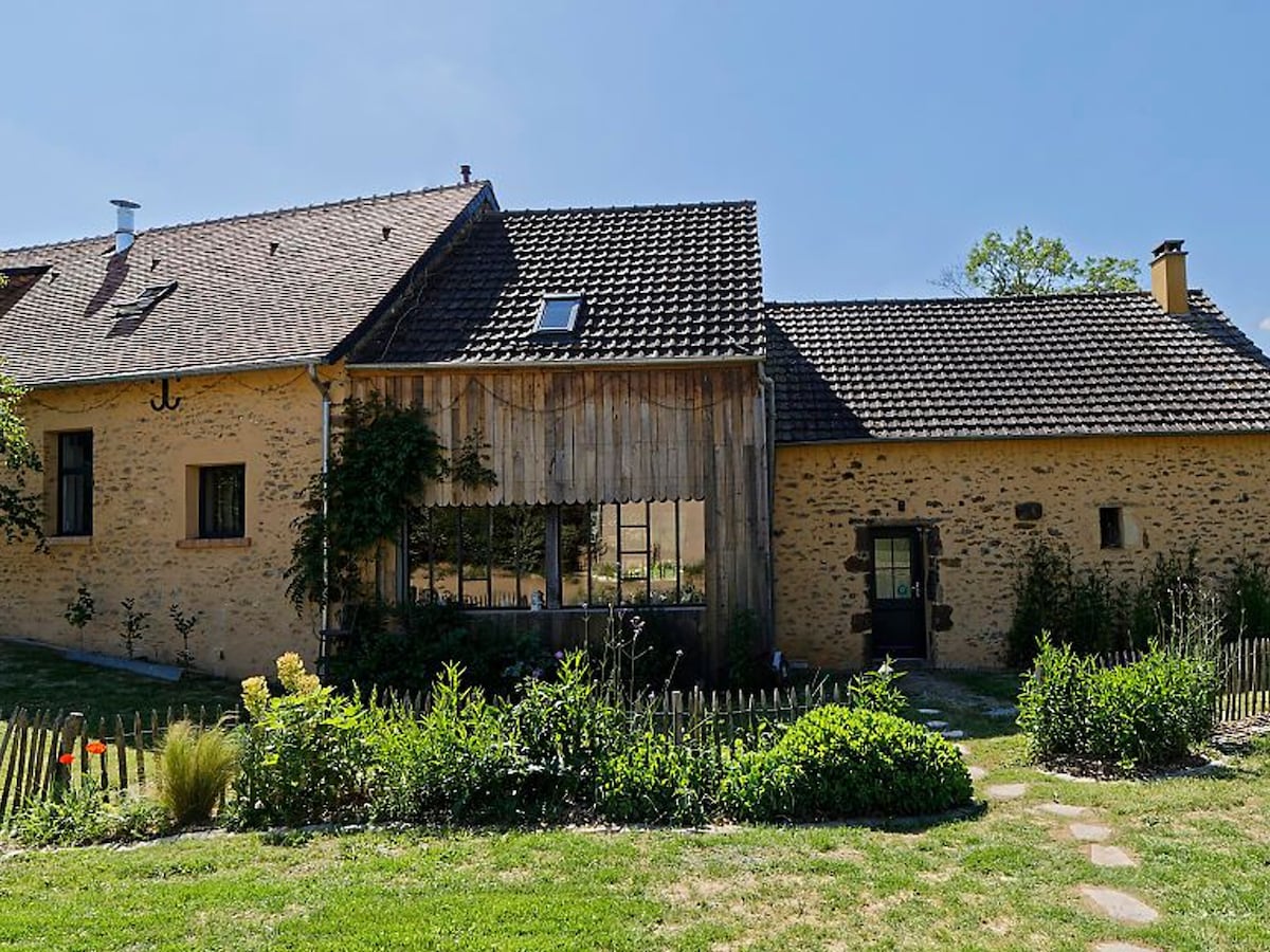 A charming farmhouse exterior is framed by a well-maintained garden, featuring various plants and flowers. The building showcases a mix of stone and wooden elements with a sloped roof. Large windows reflect natural light, enhancing the inviting ambiance of the surroundings.