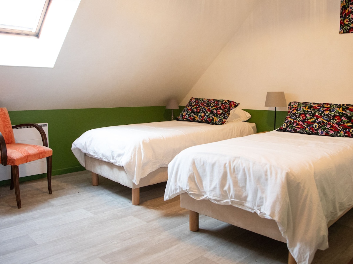 A bright room features two individual beds dressed in white linens, complemented by patterned decorative pillows. Natural light enters through a skylight, creating an airy atmosphere. An accent chair with wooden legs adds a touch of comfort to the space.