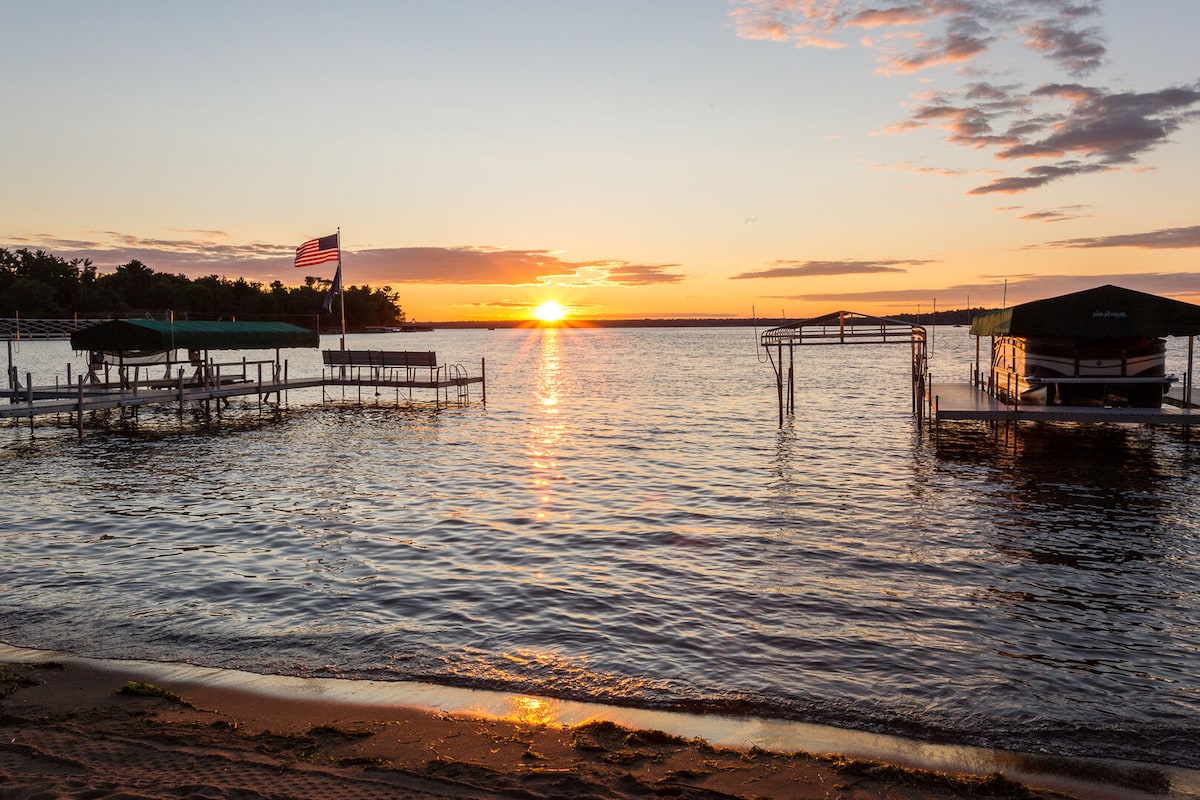 A serene lakeside scene captures the sun setting over the water, reflecting warm hues in the sky. Boat slips are visible along the shoreline, with a flag gently waving in the breeze. The sandy beach area invites relaxation beside the tranquil lake.