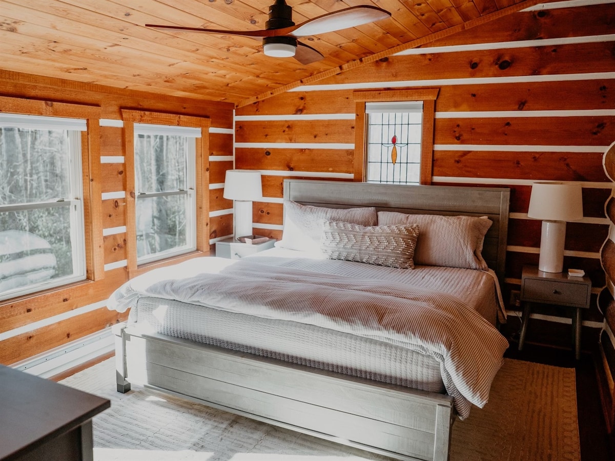 A cozy bedroom features a spacious bed with crisp white linens and decorative pillows. Two bedside tables with lamps are positioned on either side. Natural light enters through two windows, and warm wood paneling contributes to a comfortable atmosphere.