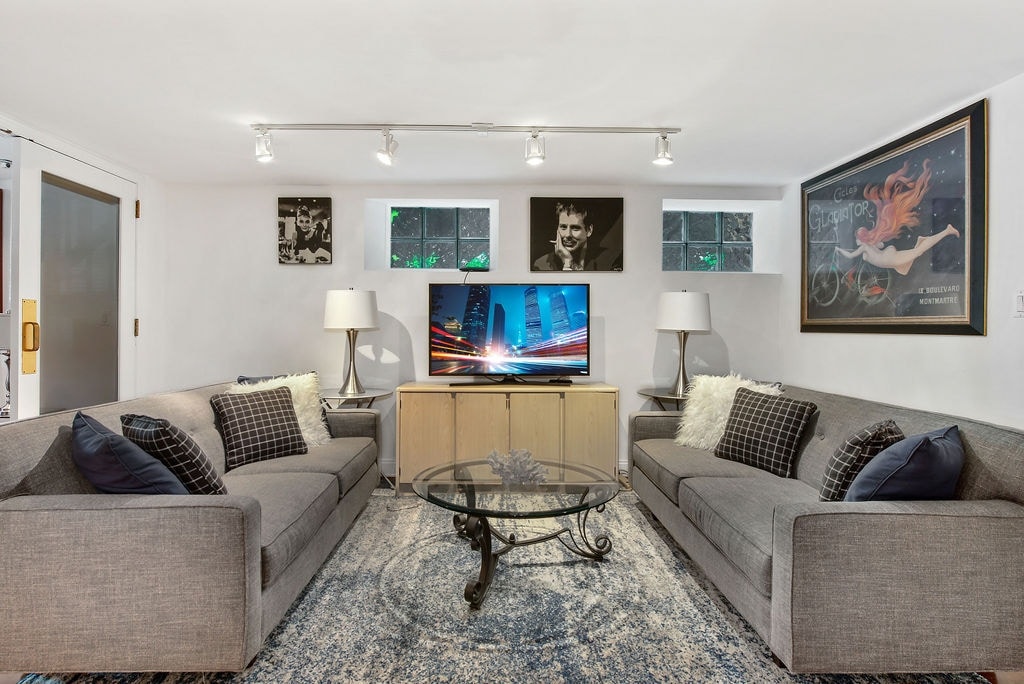 A welcoming living area features two gray sofas adorned with black and white checkered cushions. A glass-top coffee table sits at the center, complemented by two lamps flanking the furniture. Light fixtures above illuminate the space, while artwork and small windows add character to the walls.