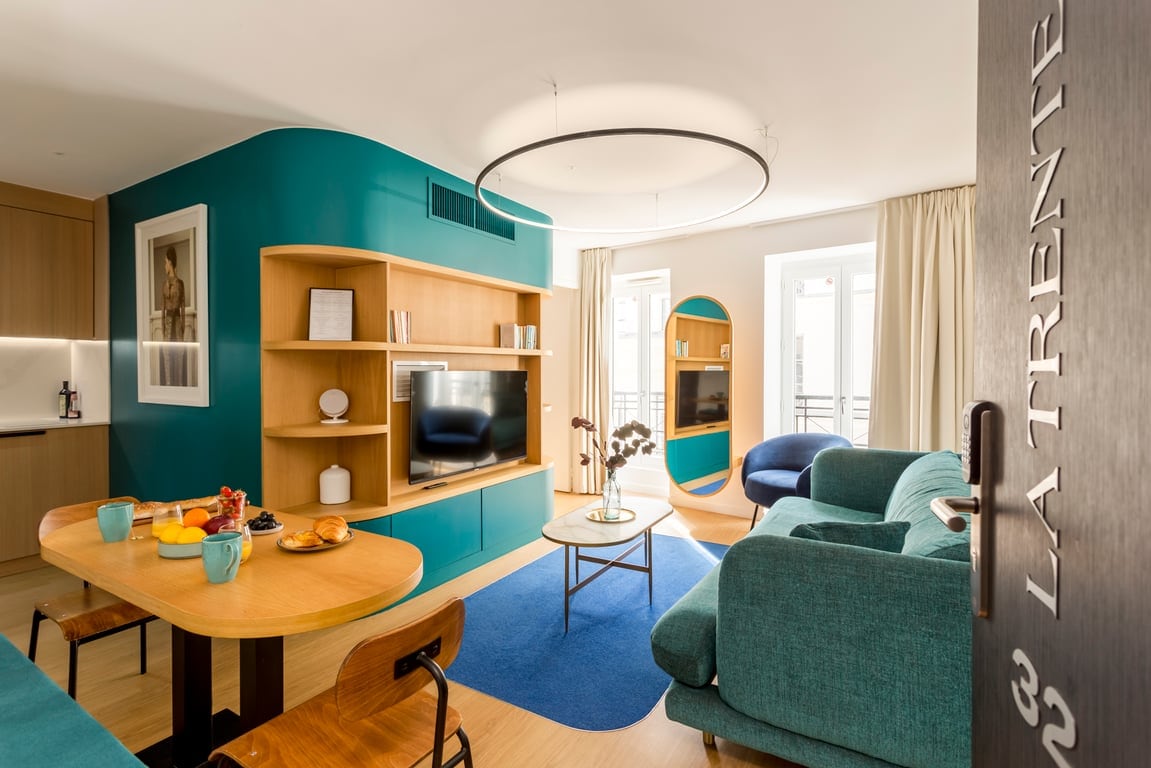 The living area features a modern design with a curved teal accent wall and wooden shelving displaying decor. A comfortable turquoise couch faces a wall-mounted TV. A round dining table is set with breakfast items, while natural light floods in from the large windows.