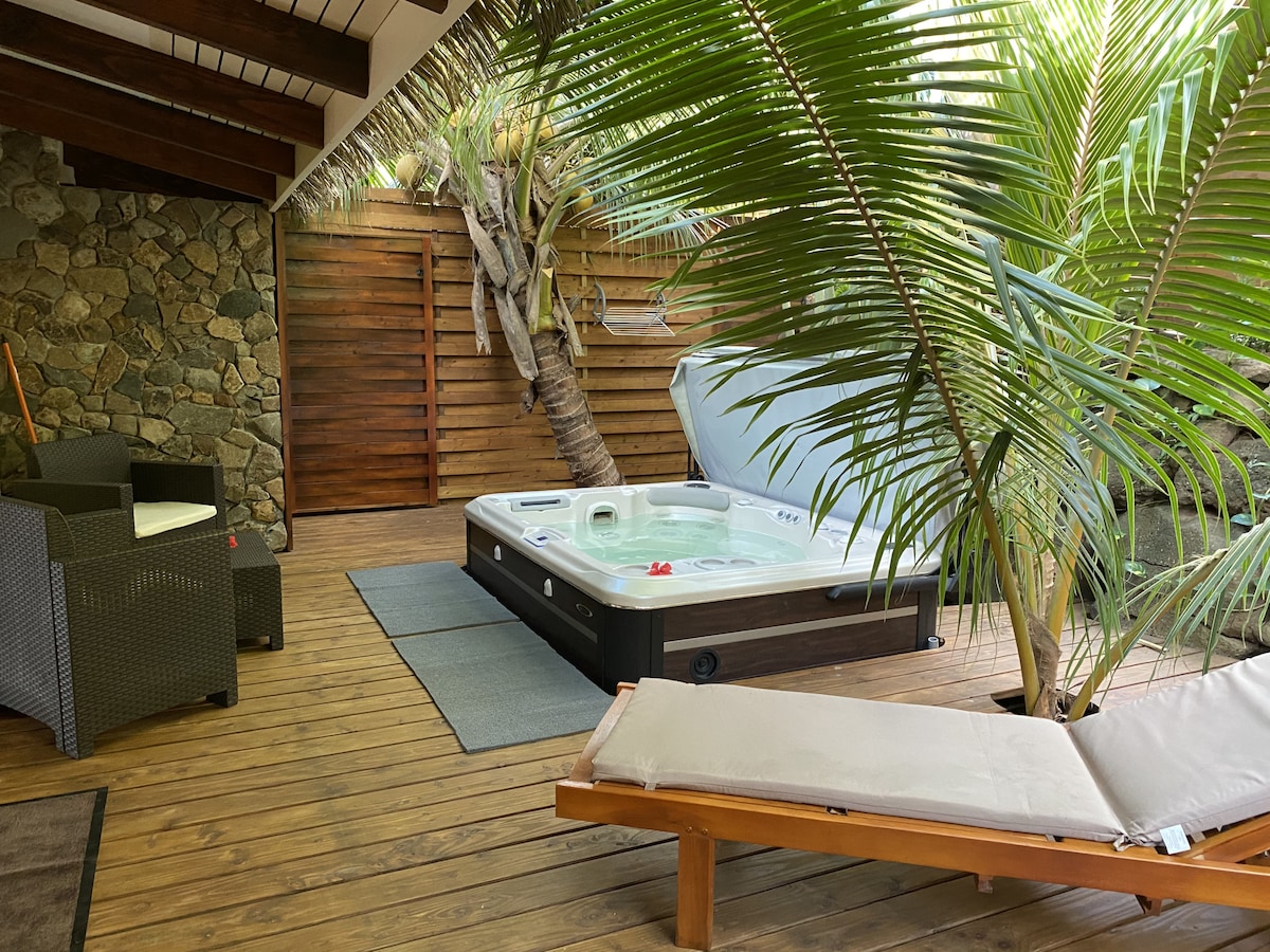 An outdoor space features a private hot tub surrounded by lush greenery and wooden decking. Comfortable seating is provided with a lounge chair and a small seating area. Natural materials create a relaxing environment, ideal for unwinding after a day at the beach.
