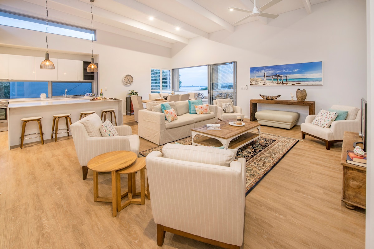A bright living area features high ceilings and large windows offering views of the exterior. Light-colored sofas and armchairs are arranged around a central coffee table. A wooden dining table in the background provides seating for six, while pendant lights hang from the ceiling.