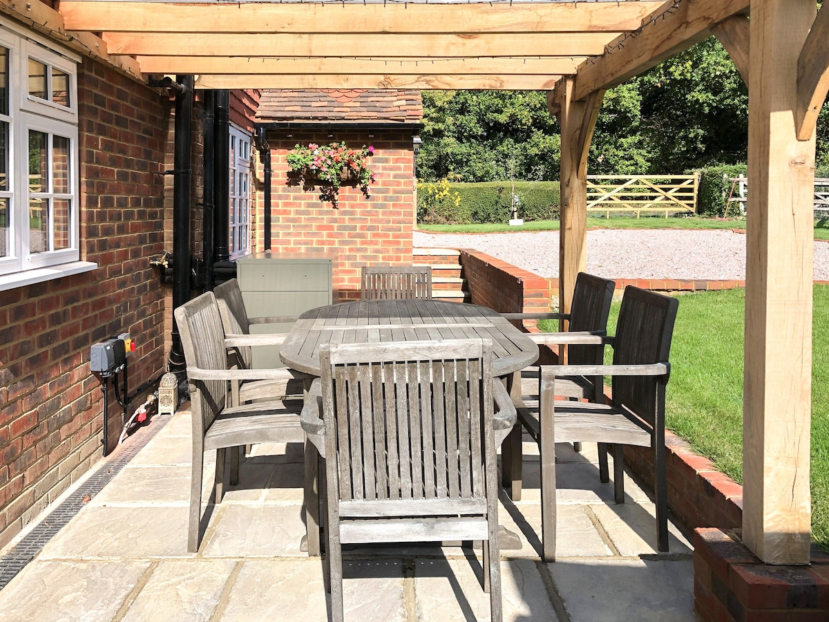 An outdoor dining area features a spacious wooden table surrounded by six matching chairs, sheltered under a wooden pergola. The patio is constructed from stone slabs, with views of the surrounding green lawn and a hedge in the background.