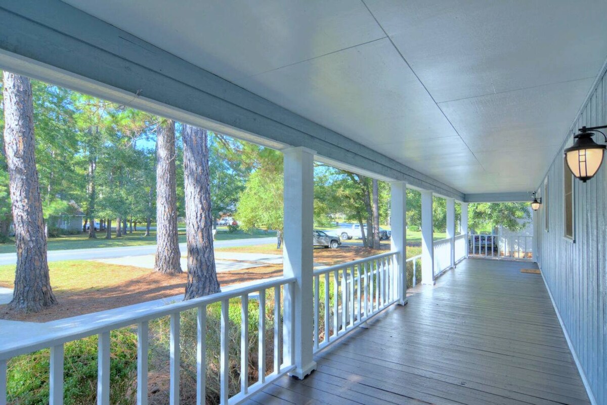 A spacious covered porch is framed by white railings and features light fixtures on either side. Sunlight streams in through large open areas, highlighting the surrounding greenery and tree-lined street beyond. The quiet setting offers a serene view of the outdoors.