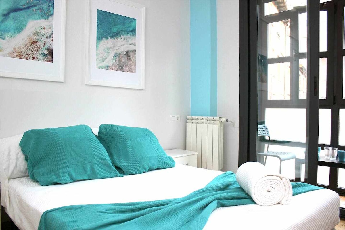 A bright sleeping area features a spacious bed adorned with two teal pillows and a rolled towel. Light floods in through large windows, with a view revealing a small outdoor seating area. Two framed artworks in shades of blue accentuate the room.