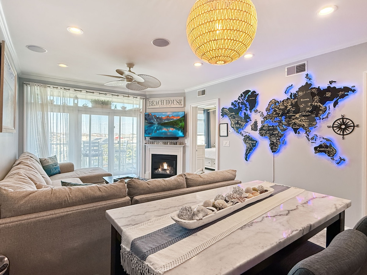 A spacious living area features a comfortable sectional sofa facing a large window with views. A stylish chandelier hangs from the ceiling, and a modern map is displayed on the wall. A decorative table with shells and a soft runner sits in the foreground.