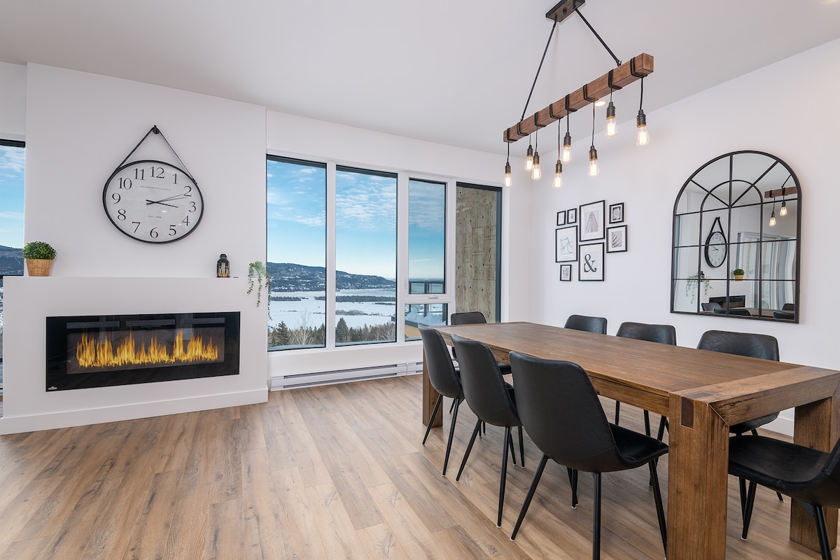 A spacious dining area features a large wooden table surrounded by sleek black chairs. A modern chandelier hangs above the table, and a decorative mirror enhances the interior. Large windows offer a scenic view of the landscape outside, while a stylish fireplace adds warmth to the space.