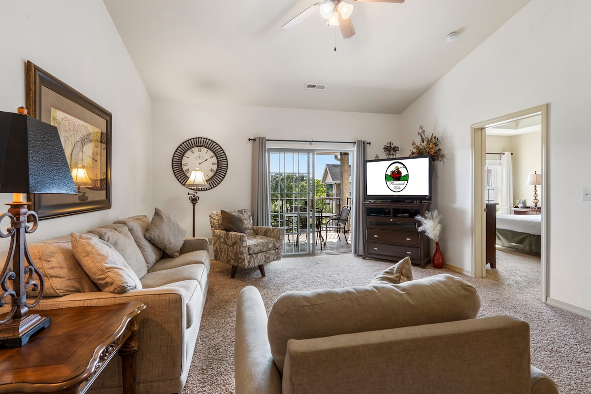 A spacious living area is depicted, featuring a comfortable sofa and a patterned armchair. A coffee table and side table are included, along with a flat-screen TV. Natural light enters through sliding glass doors leading to the private balcony, revealing views of the surroundings.